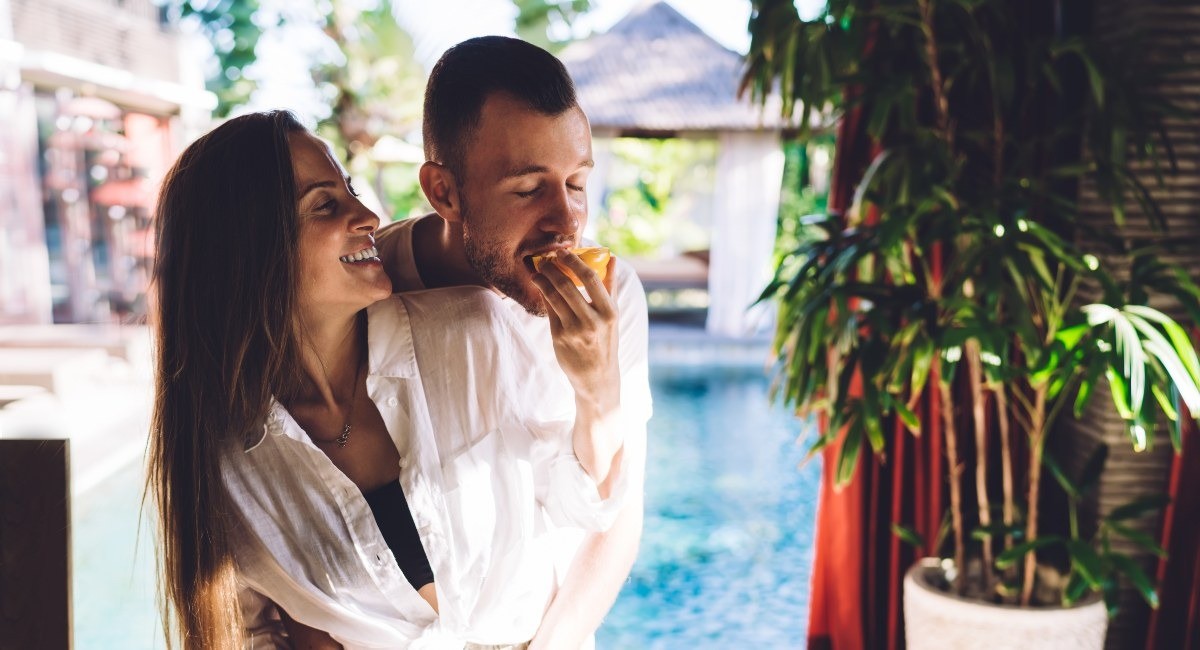 A woman with long brown hair smiles as a man with a beard eats a snack, in an affectionate embrace.