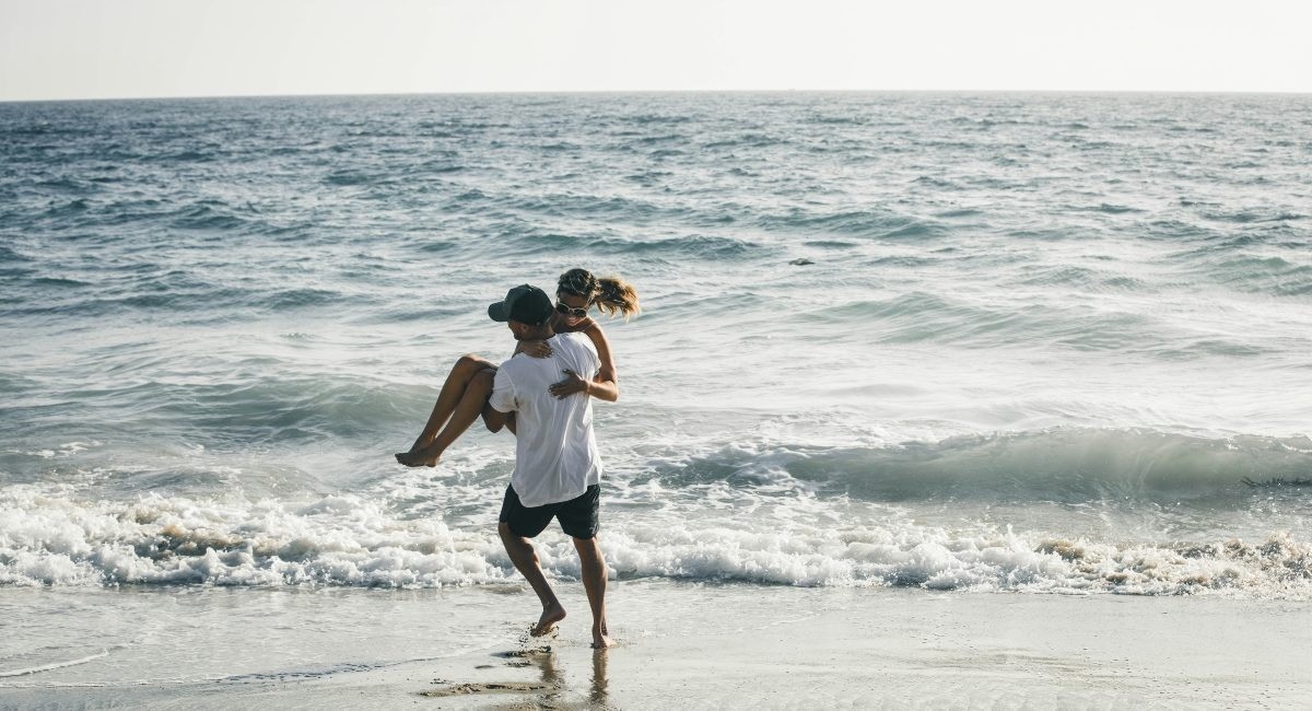A man picks up and carries a woman in his arm as waves crash against them on a beach in Grand Cayman