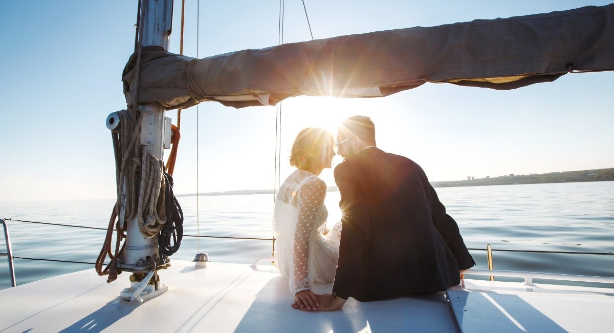 A smartly dressed couple sit on a white sailboat under a clear blue sky as the sun shines brightly behind them.