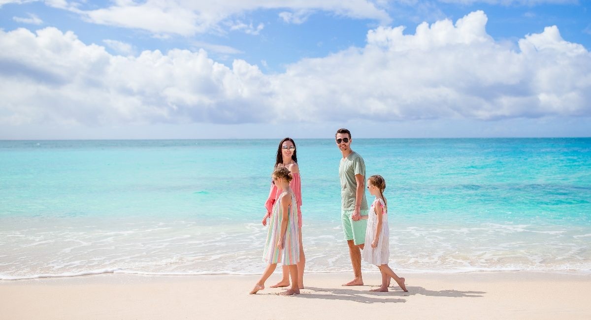 Family of four walking along a sunny tropical beach with turquoise water and white sand under a blue sky.