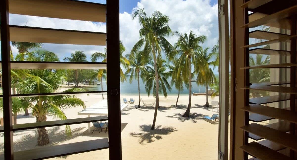 View from a window onto a tropical beach with palm trees, white sand, and lounge chairs.