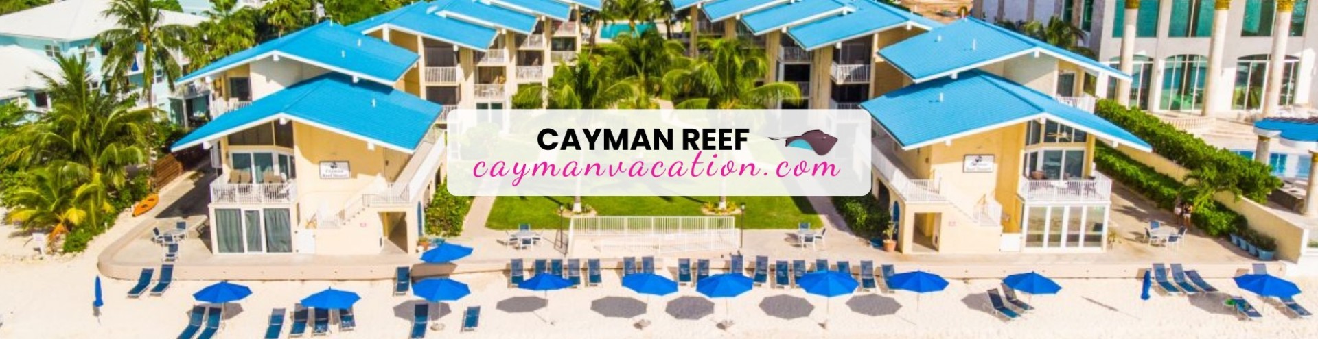 Aerial view of a beachfront resort with blue-roofed villas, palm trees, and rows of blue umbrellas and lounge chairs on the white sand.