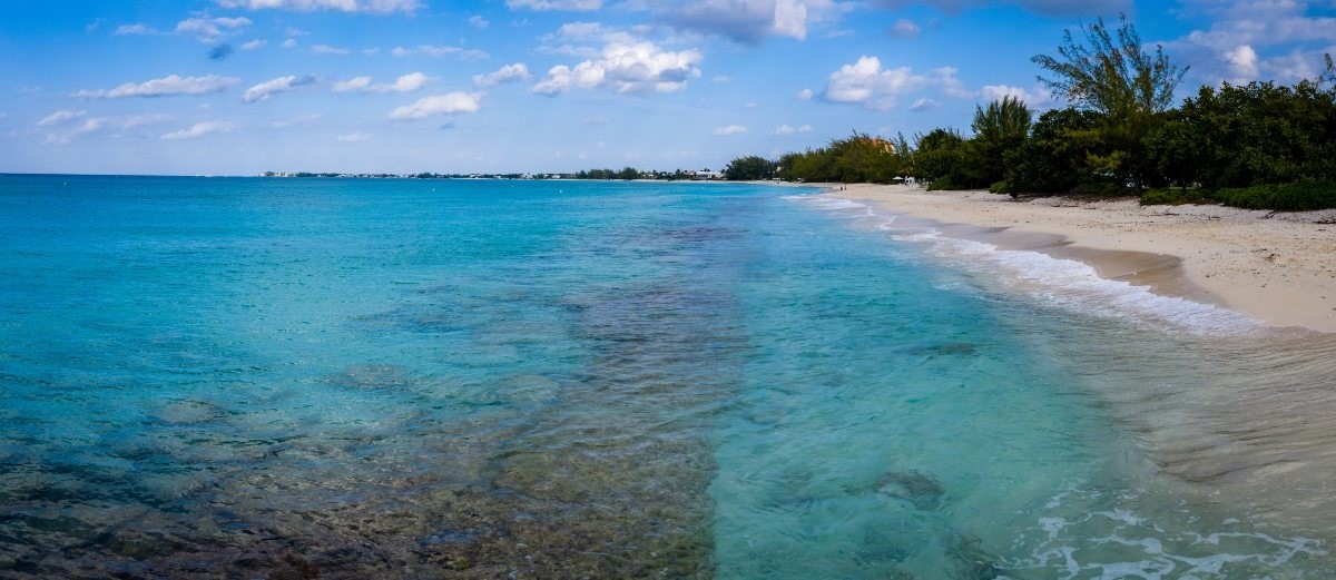 Clear turquoise sea along a white sandy beach, with green trees lining the shore under a blue sky.
