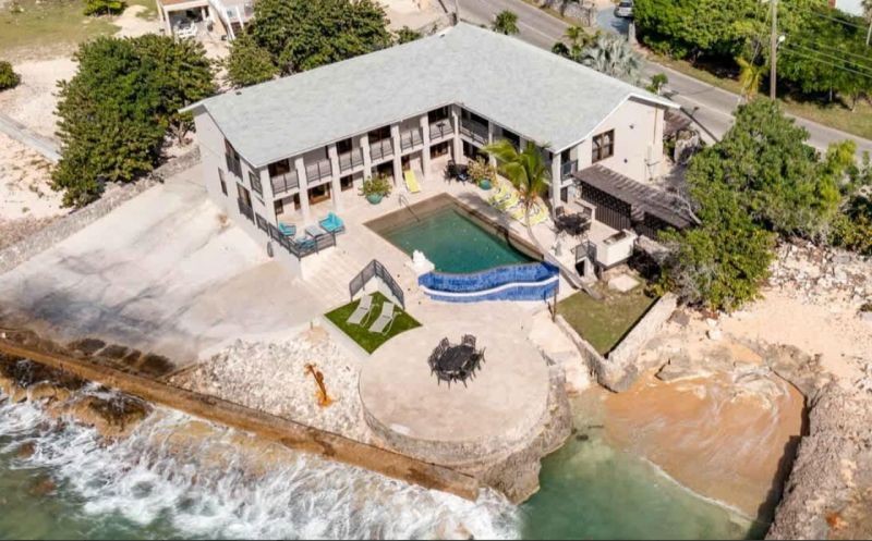 A two-story beachfront house with a swimming pool, circular patio, and seating area, beside rocky shore and turquoise water viewed from above.