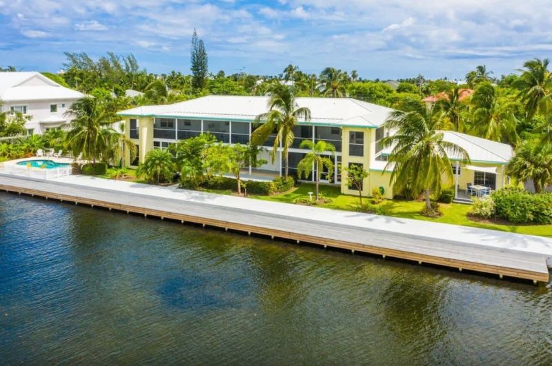 Waterfront house with a long seawall dock, palm trees, and a pool in the yard beside calm water.