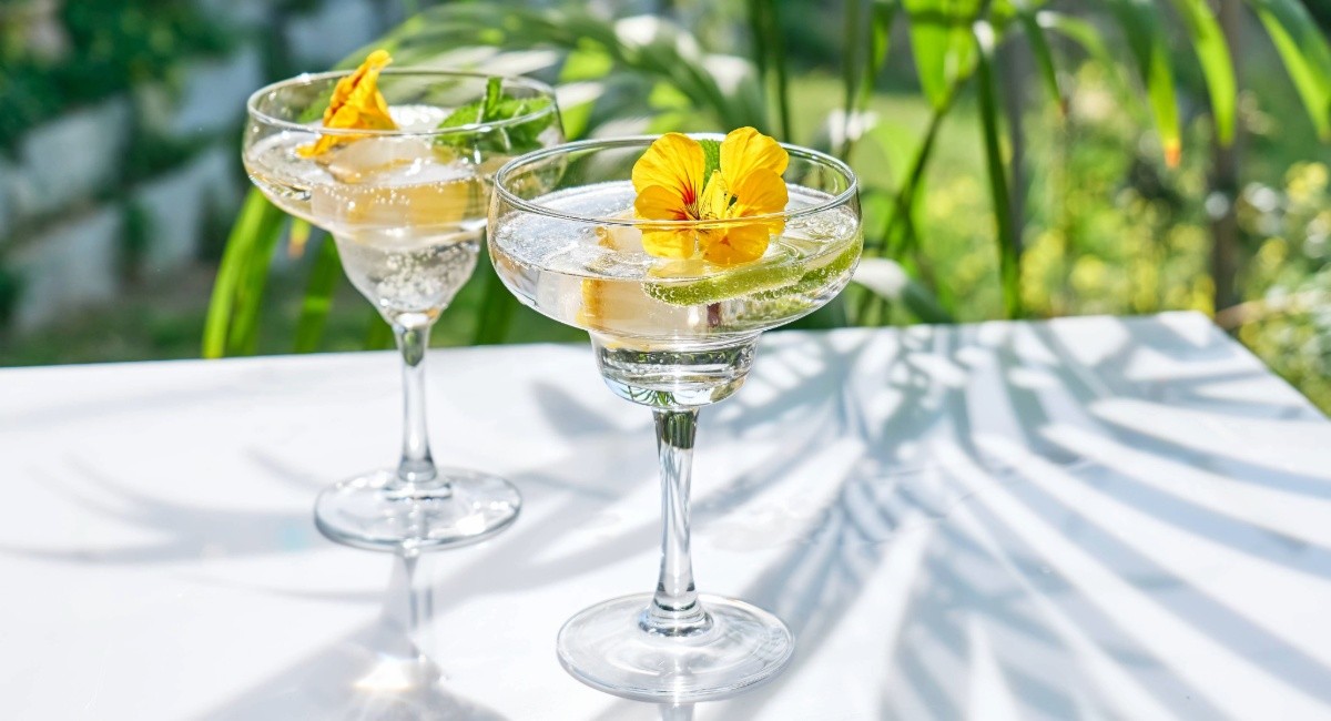 Two sparkling cocktails garnished with yellow edible flowers on a white table in palm-leaf shade