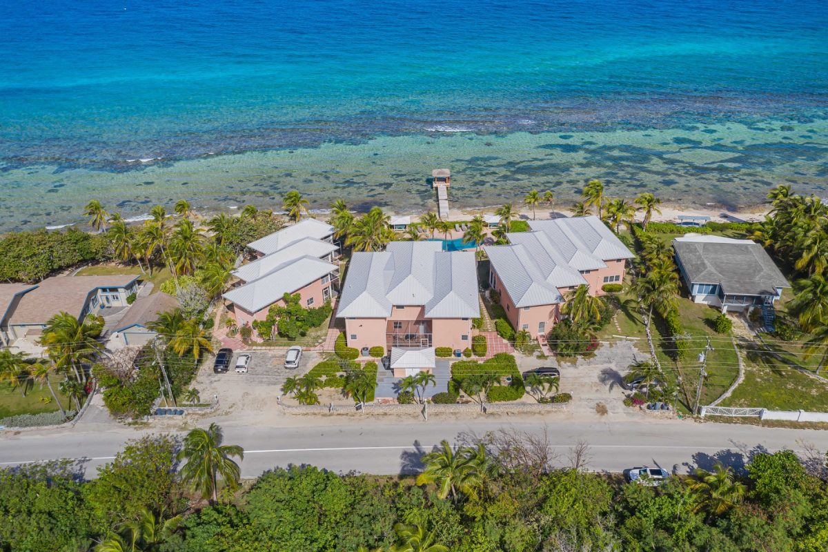 Aerial view of a coastal neighborhood with pink houses, white roofs, palm trees, and a wooden pier reaching the turquoise sea.