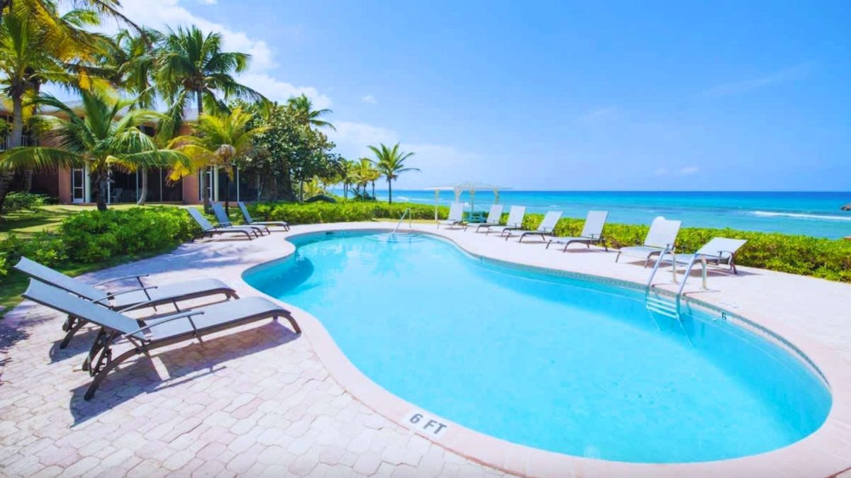 Resort pool by a tropical beach with palm trees, white lounge chairs, and bright blue water.