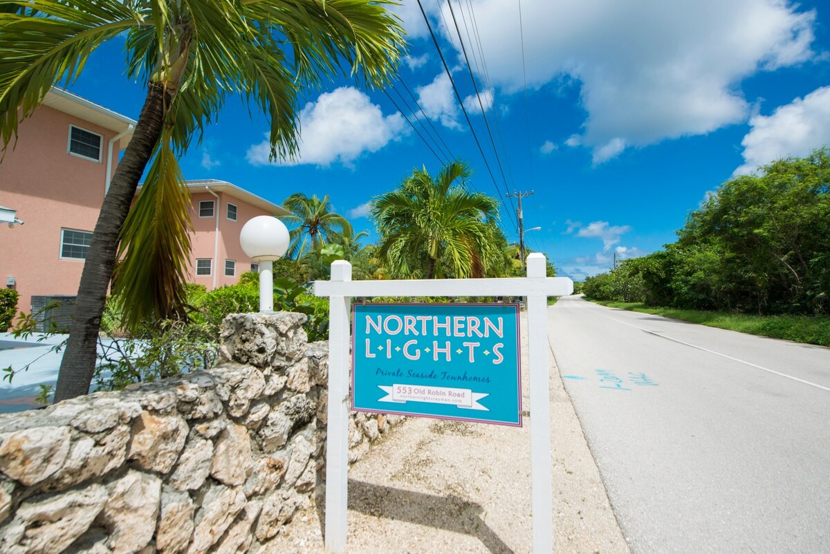 Sign reading 'Northern Lights' with 'Private Seaside Townhomes' by a white wooden frame, next to a stone wall and tropical landscaping against a blue sky.