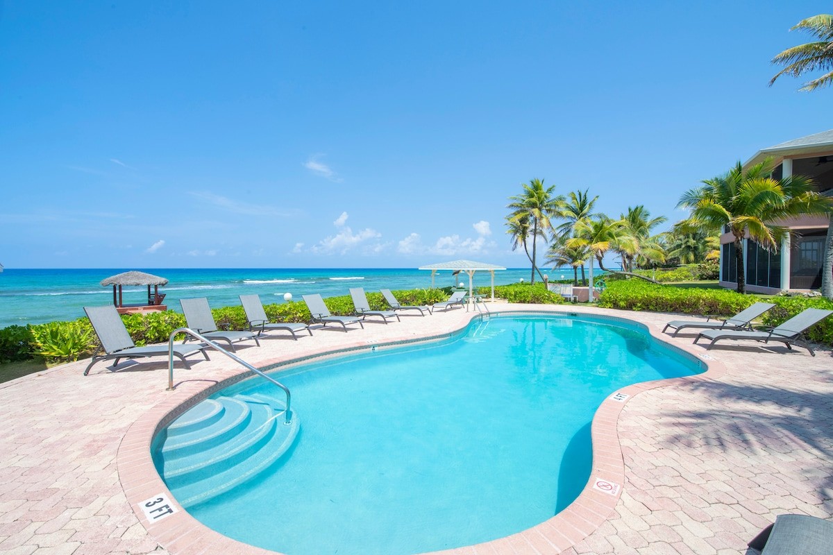 Resort pool with blue water, surrounded by lounge chairs and palm trees, overlooking the ocean on a sunny day