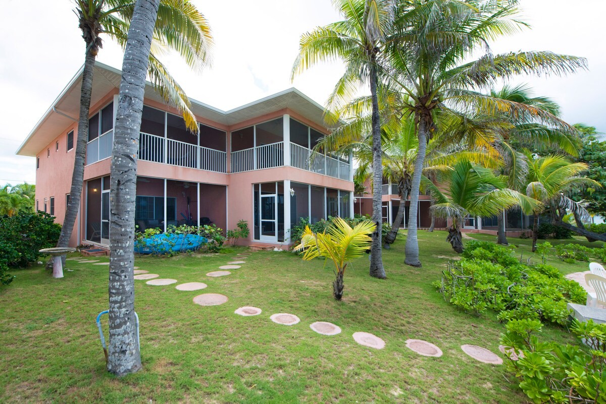 Two-story peach-colored building with screened balconies in a tropical garden, flanked by palm trees and a curved stepping-stone path in the yard, suggesting a resort villa.